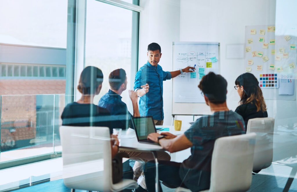 Líder apresentando informações em um quadro para a equipe durante uma reunião, ilustrando a comunicação no trabalho em equipe.